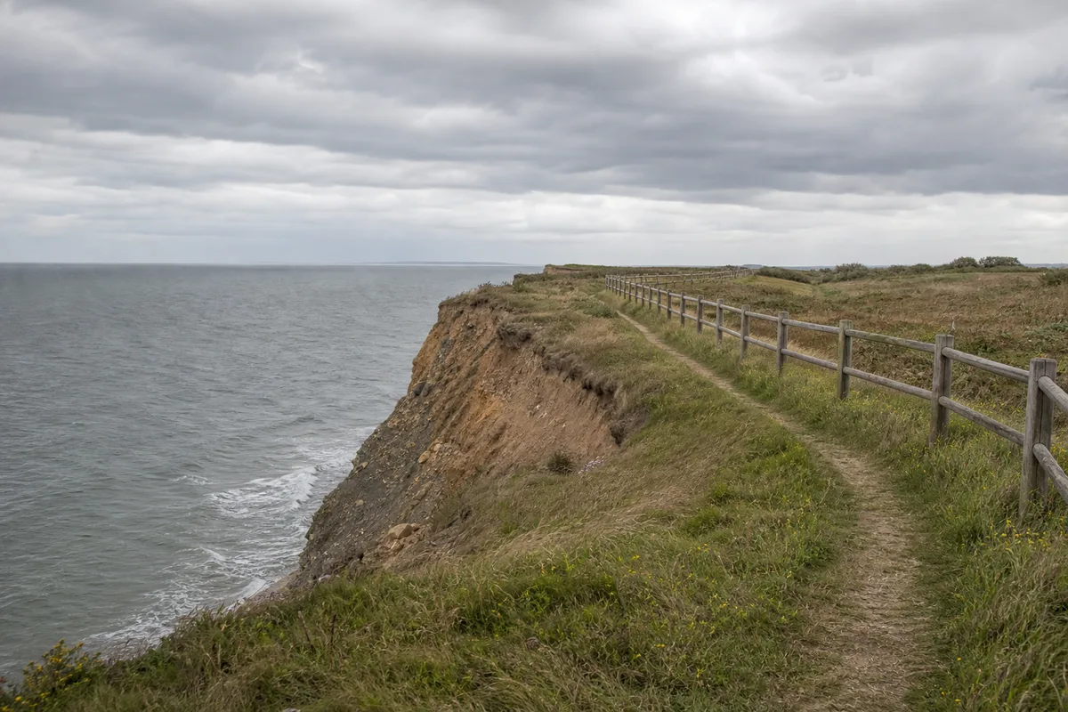Sentier côtier longeant une falaise en érosion avec clôture de sécurité au-dessus de l&rsquo;océan
